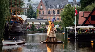 A small bonfire on a floating platform in a canal, surrounded by lush greenery and historic buildings. A boat with people is nearby. A crowd observes from a waterside cafe and a replica ship. The atmosphere is lively and festive.