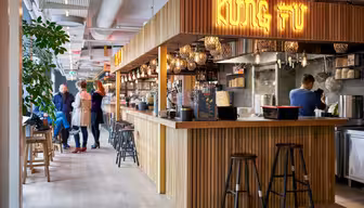A modern Asian restaurant with a wooden counter and stools, displaying a sign that reads Kung Fu. Warm lighting and decorative lamps create a cozy atmosphere. People are standing and socializing near the counter.