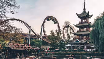 An amusement park scene with a roller coaster and riders upside-down on a loop. In the foreground, people navigate small boats on a calm pond. A tall pagoda stands to the right, surrounded by trees and blossoms.