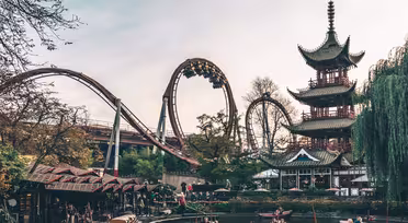 An amusement park scene with a roller coaster and riders upside-down on a loop. In the foreground, people navigate small boats on a calm pond. A tall pagoda stands to the right, surrounded by trees and blossoms.