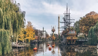 A scenic view of a theme park lake with an old ship replica, surrounded by autumn-colored trees. Floating nearby are small orange buoys. In the background, a tall tower rises against a blue sky.
