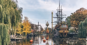 A scenic view of a theme park lake with an old ship replica, surrounded by autumn-colored trees. Floating nearby are small orange buoys. In the background, a tall tower rises against a blue sky.