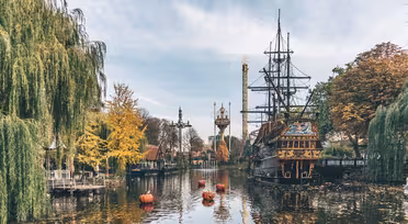 A scenic view of a theme park lake with an old ship replica, surrounded by autumn-colored trees. Floating nearby are small orange buoys. In the background, a tall tower rises against a blue sky.