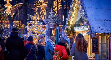 People gather around a festive market stall adorned with lights and snow, in a winter setting. Warm clothing, including coats and scarves, suggest the cold weather. In the background, more lights decorate trees, creating a cozy, festive atmosphere.
