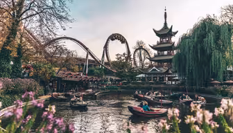 A picturesque amusement park scene with boats on a pond, a looping roller coaster, and a traditional pagoda surrounded by lush greenery and flowering plants. The sky is clear, adding to the vibrant and lively atmosphere.