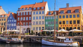 Colorful, historic buildings line the Nyhavn waterfront in Copenhagen, Denmark. Docked boats float gently in the canal, with outdoor cafes and a sunny blue sky completing the lively scene.
