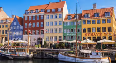Colorful, historic buildings line the Nyhavn waterfront in Copenhagen, Denmark. Docked boats float gently in the canal, with outdoor cafes and a sunny blue sky completing the lively scene.