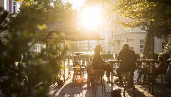 People sit at outdoor cafe tables under umbrellas on a sunlit street. The sun shines through the trees, casting long shadows. The atmosphere is relaxed and the surroundings suggest a warm afternoon.