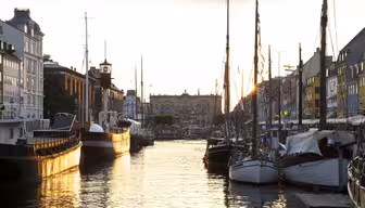 A serene canal lined with boats in the evening, reflecting the golden sunlight. Historic buildings with traditional architecture flank both sides. The atmosphere is calm and picturesque.