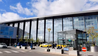 Exterior of a modern glass airport terminal with a blue sky above. Yellow taxis and people are visible near the entrance. A pedestrian crossing and no entry sign are in the foreground.