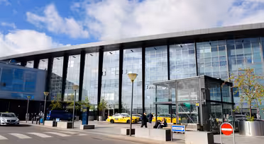 Exterior of a modern glass airport terminal with a blue sky above. Yellow taxis and people are visible near the entrance. A pedestrian crossing and no entry sign are in the foreground.
