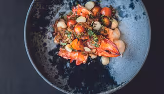 A dish of vibrant seafood salad featuring lobster claws, cherry tomatoes, white beans, and fresh herbs, served in a dark, rustic bowl against a black background.