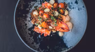 A dish of vibrant seafood salad featuring lobster claws, cherry tomatoes, white beans, and fresh herbs, served in a dark, rustic bowl against a black background.