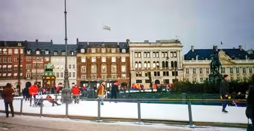 People ice skate on an outdoor rink in front of historic European-style buildings. Some wear red jackets, and a green statue is visible in the background. A clear sky suggests daytime in a lively urban setting.