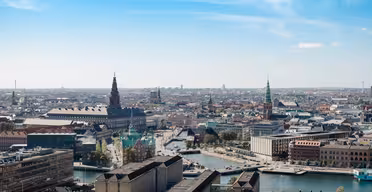 Aerial view of a European cityscape featuring a mix of historic and modern buildings, a river with several bridges, and church spires under a clear blue sky.