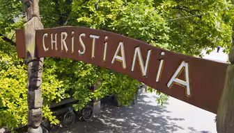 A wooden sign with the word Christiania in yellow capital letters arches over a pathway bordered by lush green trees.Several bicycles are parked beneath the sign. Totem-like carvings decorate the poles supporting the sign.