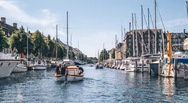 A scenic canal with several boats on a sunny day. The water is calm, and sailboats are docked along both sides of the canal. Buildings line the waterfront, and trees add greenery to the scene under a clear blue sky.