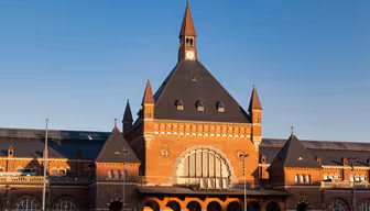 A red brick train station with a large arched window and a central clock tower under a blue sky. The building features pointed roofs and decorative elements, with multiple smaller towers on either side.