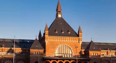A red brick train station with a large arched window and a central clock tower under a blue sky. The building features pointed roofs and decorative elements, with multiple smaller towers on either side.