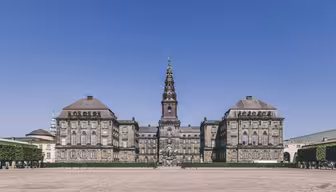 The image shows a large, historic European building with a central tower topped by a spire. The facade is symmetrical, with a courtyard in front. The sky is clear and blue, and there are trimmed hedges lining the sides.