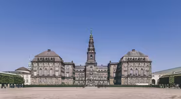The image shows a large, historic European building with a central tower topped by a spire. The facade is symmetrical, with a courtyard in front. The sky is clear and blue, and there are trimmed hedges lining the sides.