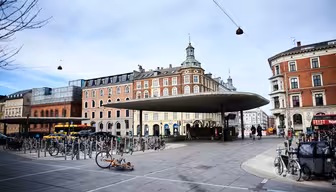 Urban scene with a circular bus stop shelter surrounded by many parked bicycles. Historic buildings with ornate facades are in the background under a cloudy sky. Few pedestrians and a parked bus are visible.