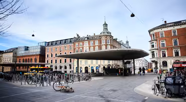 Urban scene with a circular bus stop shelter surrounded by many parked bicycles. Historic buildings with ornate facades are in the background under a cloudy sky. Few pedestrians and a parked bus are visible.