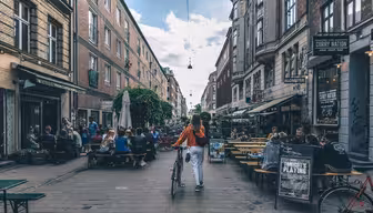 A person in an orange shirt walks a bicycle down a lively street. People sit at outdoor cafes on both sides, enjoying the atmosphere. The street is lined with brick buildings under a partly cloudy sky.