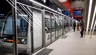 A modern train stopped at an underground station with glass doors separating the platform. Passengers are visible inside and a few people stand on the platform. Illuminated signs and sleek design elements are present.