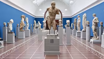 A museum gallery showcasing various ancient stone sculptures on pedestals. The room has a blue wall and a high arched ceiling with skylights, creating a bright and airy atmosphere. Visitors are viewing the sculptures.