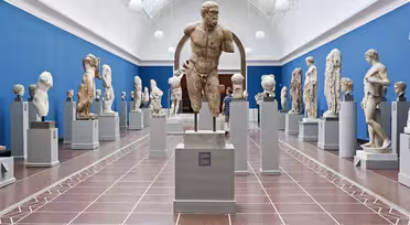 A museum gallery showcasing various ancient stone sculptures on pedestals. The room has a blue wall and a high arched ceiling with skylights, creating a bright and airy atmosphere. Visitors are viewing the sculptures.