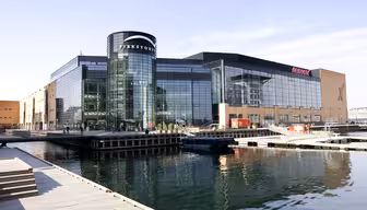 Large modern glass building beside a calm body of water, featuring the name Fisketorvet prominently. The structure has a mix of glass and brick elements, and the scene includes reflections on the water and a clear blue sky.