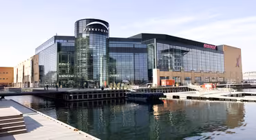Large modern glass building beside a calm body of water, featuring the name Fisketorvet prominently. The structure has a mix of glass and brick elements, and the scene includes reflections on the water and a clear blue sky.