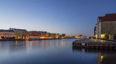 Waterfront view of a city at dusk, showcasing calm waters and buildings with illuminated windows. The sky is a gradient from light to deep blue, and the scene reflects on the water, creating a peaceful and serene atmosphere.