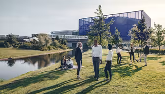 A group of people stand and converse on a grassy area near a pond. In the background, a modern building with glass and metal structures reflects the sunlight. Some individuals are sitting on the grass, while others are walking nearby.