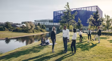 A group of people stand and converse on a grassy area near a pond. In the background, a modern building with glass and metal structures reflects the sunlight. Some individuals are sitting on the grass, while others are walking nearby.