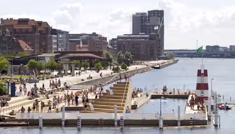 A lively waterfront scene featuring a floating swimming pool surrounded by people on a sunny day. Modern and historic buildings line the promenade, with a green flag flying atop a distinctive white and red tower in the pool area.