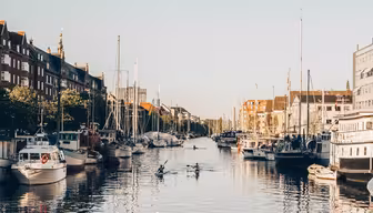 A calm canal scene with several small boats docked along both sides. Three kayakers paddle through the water. Buildings line the canal under a clear blue sky, adding a serene urban backdrop to the setting.