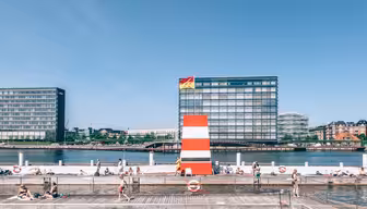 People swimming and sunbathing at a waterfront area with a wooden deck. A tall, striped structure stands at the waters edge. Modern buildings and a clear blue sky are in the background.