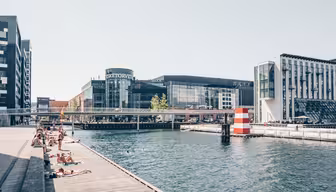 People sunbathe on wooden decks next to a canal with clear water. Modern buildings, including a curved glass structure, line the opposite side. A pedestrian bridge spans the canal, and a red and white striped cylinder adds nautical charm.