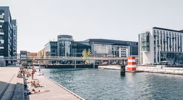 People sunbathe on wooden decks next to a canal with clear water. Modern buildings, including a curved glass structure, line the opposite side. A pedestrian bridge spans the canal, and a red and white striped cylinder adds nautical charm.