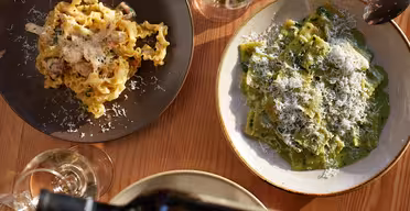 Two plates of pasta on a wooden table: one with curly, ribbon-like pasta and the other with green-filled ravioli, both topped with grated cheese. A hand pours wine into a glass, with additional wine glasses nearby.