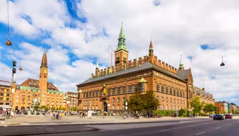 A vibrant scene of Copenhagens City Hall, featuring its red-brick architecture and green spires under a partly cloudy sky. People are walking in the foreground near a fountain, and the bustling street adds life to the historic setting.