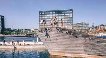 People dive and sit on a wooden ramp by the water, with modern buildings in the background. The sunny day shows a lively, recreational scene by the waters edge.