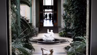 See through an arched doorway into a grand interior with lush plants, a central fountain, and a classical statue in the foreground. Two distant figures walk past the entrance in the background, framed by columns.