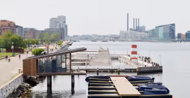 Waterfront scene with a modern wooden pavilion and small docked boats in the foreground. People stroll along a waterside path lined with greenery. In the background, a large industrial building and modern architecture are visible along the water.