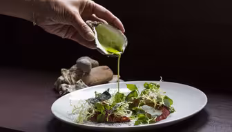 A hand holds a half shell, pouring bright green sauce over a plate of salad with leafy greens and thinly sliced meat. Oyster shells rest in the background on a dark surface.