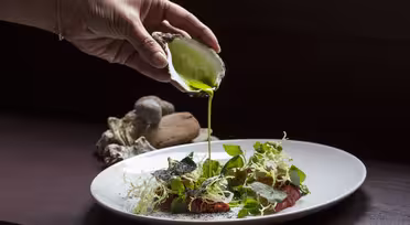 A hand holds a half shell, pouring bright green sauce over a plate of salad with leafy greens and thinly sliced meat. Oyster shells rest in the background on a dark surface.