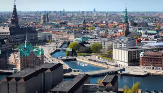 Aerial view of a cityscape featuring a canal winding through historic buildings with spires and towers. The scene includes greenery and modern structures, set under a clear blue sky.