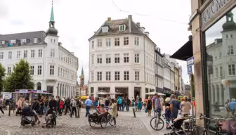 A bustling European city street with people walking, cycling, and sitting at an outdoor cafe. The scene features cobblestone pavement, historic buildings with pointed rooftops, and a mix of pedestrians including parents with strollers.